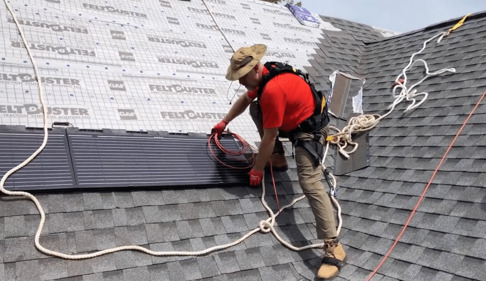 roofer installs flexible solar panels on a shingled roof while secured with safety ropes and wearing protective climbing equipment and gear
