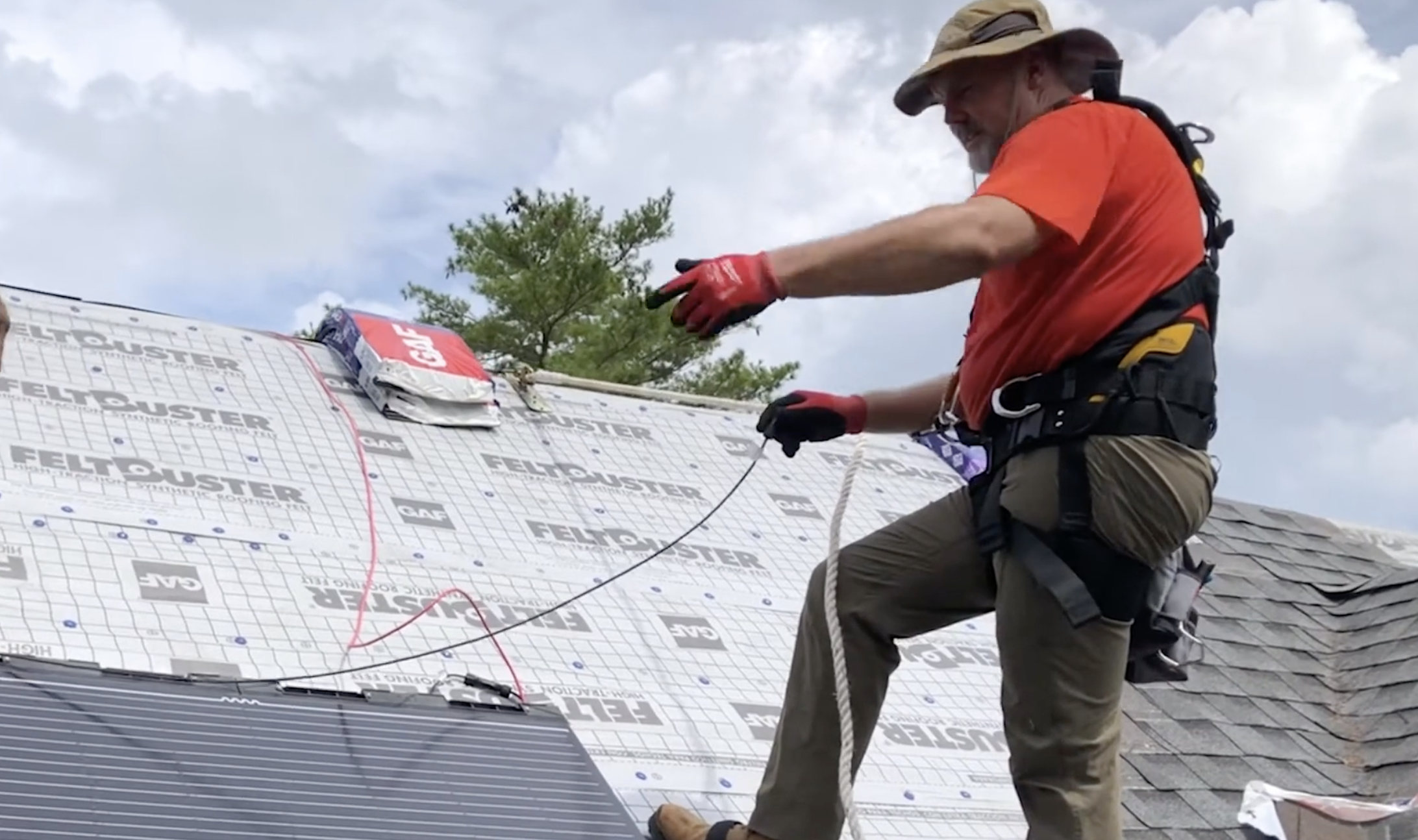 roofer wearing a safety harness connects wiring beside flexible solar panels on a partially shingled residential roof under cloudy skies
