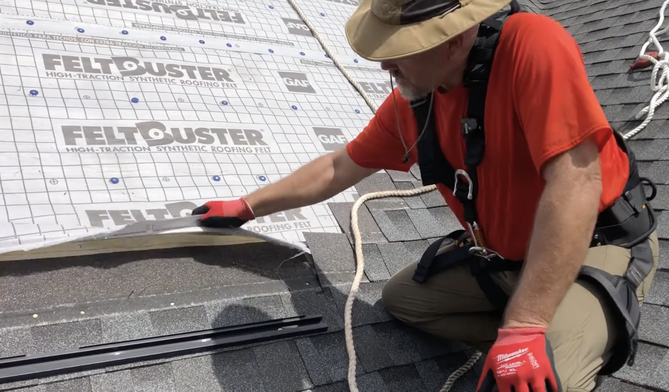 roofer wearing safety gear lifts roofing underlayment beside mounting rails while preparing a shingled roof for solar panel installation work