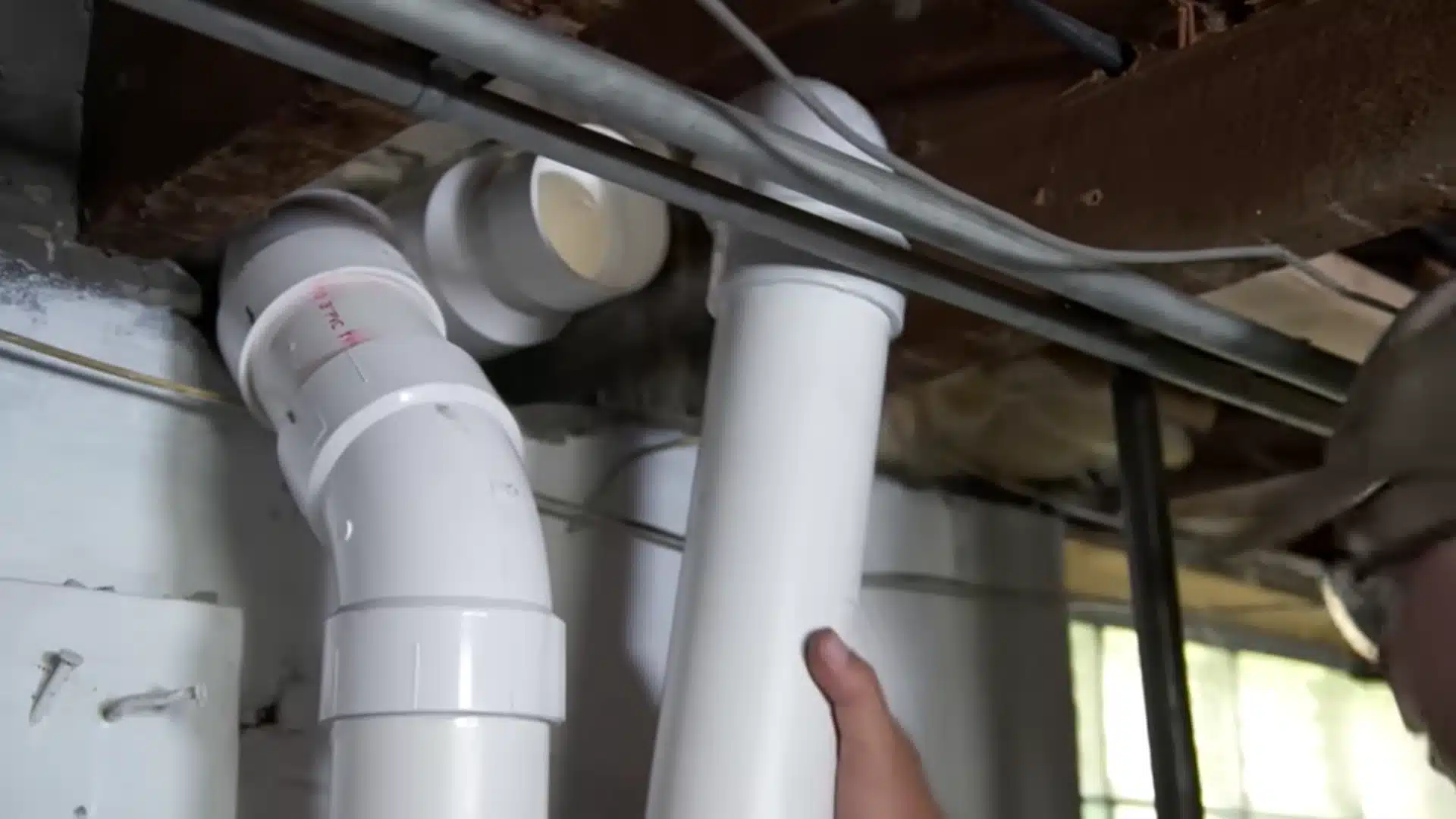 Worker connects white PVC intake and exhaust vent pipes beneath an open basement ceiling